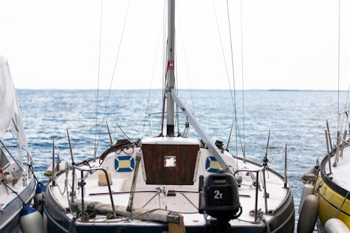 A sailboat is docked at a marina, surrounded by calm blue waters under a bright sky. The boat is equipped with various nautical instruments and ropes, with a wooden cabin door and a number of fenders attached to its sides.