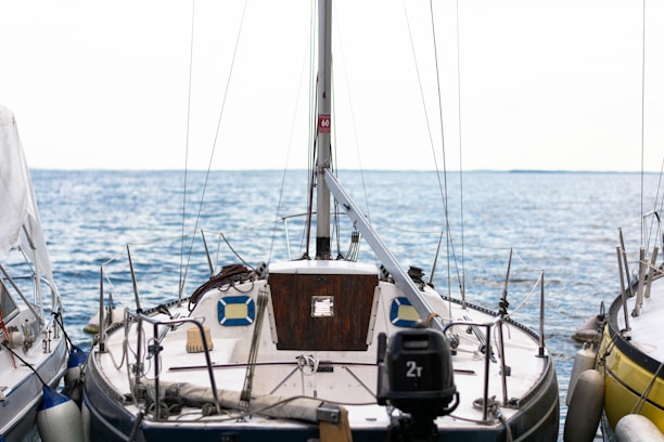 A sailboat is docked at a marina, surrounded by calm blue waters under a bright sky. The boat is equipped with various nautical instruments and ropes, with a wooden cabin door and a number of fenders attached to its sides.