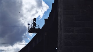 A silhouette of a structure is visible against a cloudy sky. The structure appears to be an elevated platform with a railing and possibly some equipment or machinery. There's also a small bird flying nearby.