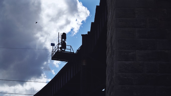A silhouette of a structure is visible against a cloudy sky. The structure appears to be an elevated platform with a railing and possibly some equipment or machinery. There's also a small bird flying nearby.