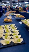 An assortment of small, meticulously arranged appetizers is displayed on a large table. The appetizers include green and white balls in wooden bowls, pastries, and dishes topped with creamy sauces. People in the background are gathered, possibly for an event, with some picking up food items.