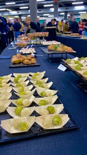 An assortment of small, meticulously arranged appetizers is displayed on a large table. The appetizers include green and white balls in wooden bowls, pastries, and dishes topped with creamy sauces. People in the background are gathered, possibly for an event, with some picking up food items.