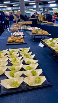 An assortment of small, meticulously arranged appetizers is displayed on a large table. The appetizers include green and white balls in wooden bowls, pastries, and dishes topped with creamy sauces. People in the background are gathered, possibly for an event, with some picking up food items.