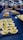 An assortment of small, meticulously arranged appetizers is displayed on a large table. The appetizers include green and white balls in wooden bowls, pastries, and dishes topped with creamy sauces. People in the background are gathered, possibly for an event, with some picking up food items.