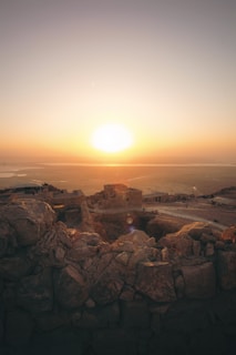 A sunrise casts a warm glow over an ancient, rocky fortress on a hilltop, with expansive desert landscapes stretching into the horizon. The scene conveys tranquility and timelessness, as the first light softly touches the rugged stone structures and walls.