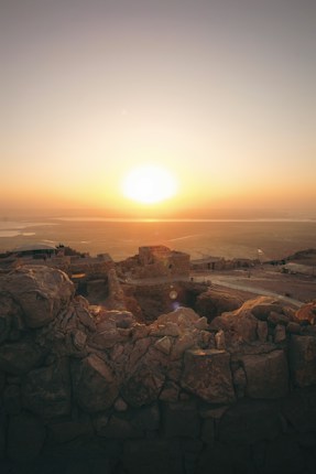 A sunrise casts a warm glow over an ancient, rocky fortress on a hilltop, with expansive desert landscapes stretching into the horizon. The scene conveys tranquility and timelessness, as the first light softly touches the rugged stone structures and walls.