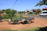 A rural roadside scene with a motorcycle attached to a small trailer parked on a dirt path. There is another motorcycle in the background and a white minivan driving on the paved road. Residential houses and a variety of green bushes and trees are visible along the roadside. Overhead power lines are strung across the sky, which is clear and blue. The ground is a mix of dirt and sparse grass.