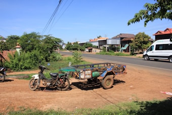 A rural roadside scene with a motorcycle attached to a small trailer parked on a dirt path. There is another motorcycle in the background and a white minivan driving on the paved road. Residential houses and a variety of green bushes and trees are visible along the roadside. Overhead power lines are strung across the sky, which is clear and blue. The ground is a mix of dirt and sparse grass.