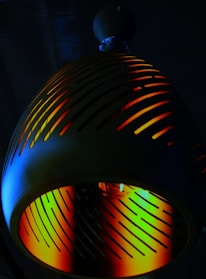 A dimly lit decorative lamp with a spherical top and a slotted, dome-shaped shade. Vibrant colors such as orange, yellow, green, and blue emit through the slots, creating a multicolored glow.