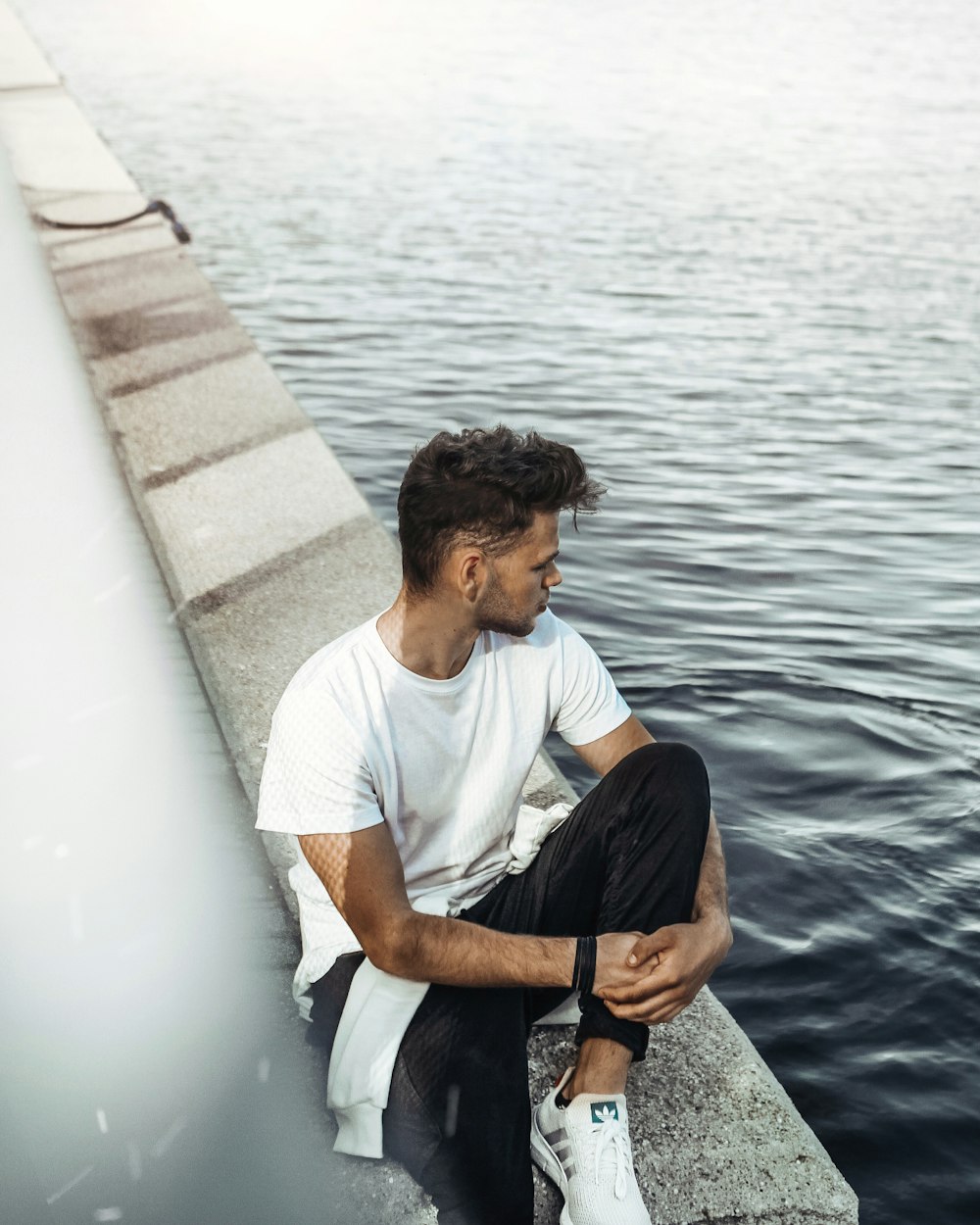man in white crew neck t-shirt and black pants sitting on concrete dock during daytime
