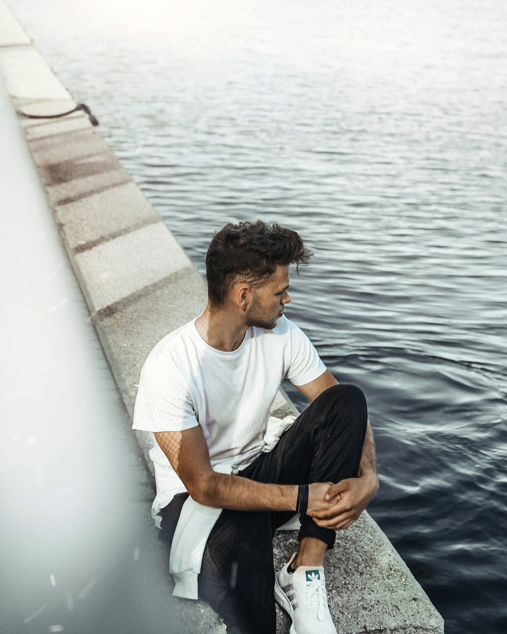 Mann in weißem Rundhals-T-Shirt und schwarzer Hose, der tagsüber auf dem Betondock sitzt