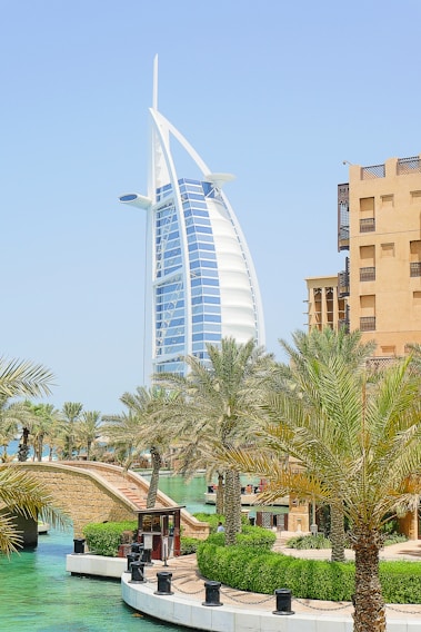 A futuristic, sail-shaped skyscraper dominates the skyline, surrounded by lush palm trees and a serene canal. Traditional architecture buildings with beige exteriors stand nearby, blending modern and traditional elements.