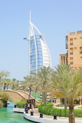 A futuristic, sail-shaped skyscraper dominates the skyline, surrounded by lush palm trees and a serene canal. Traditional architecture buildings with beige exteriors stand nearby, blending modern and traditional elements.