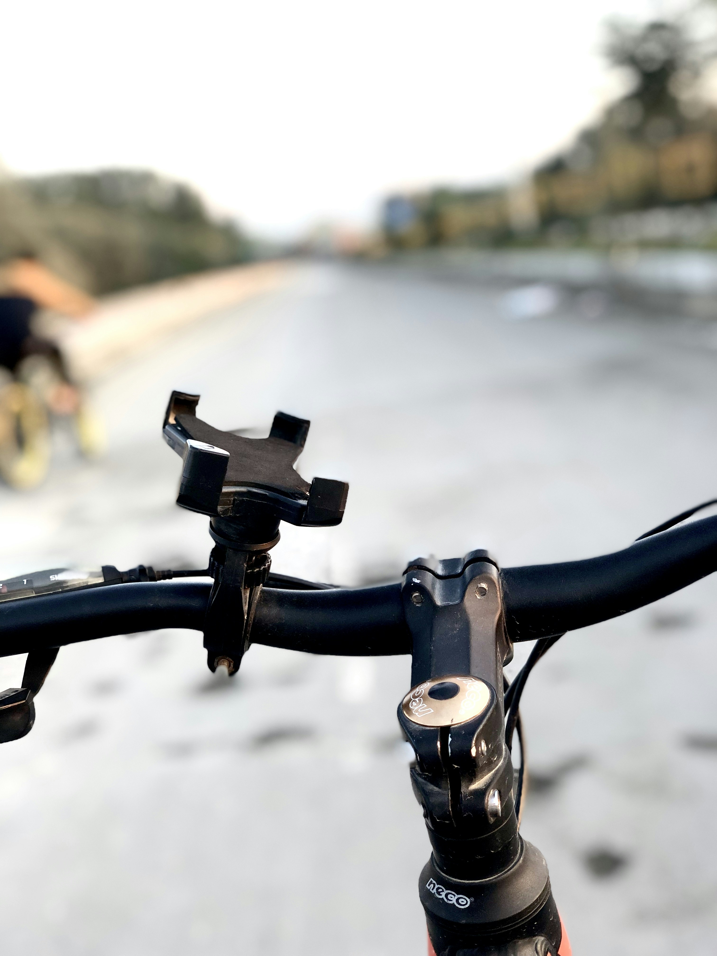Empty black four-corner claw phone holder clamped to a bicycle handlebar beside the stem, a riverside path blurred behind