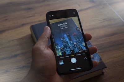 A hand is holding a smartphone, displaying a book cover image titled 'Life 3.0' by Max Tegmark, with a city skyline featured prominently. The background shows a wooden surface with a closed book.