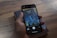 A hand is holding a smartphone, displaying a book cover image titled 'Life 3.0' by Max Tegmark, with a city skyline featured prominently. The background shows a wooden surface with a closed book.