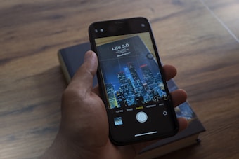A hand is holding a smartphone, displaying a book cover image titled 'Life 3.0' by Max Tegmark, with a city skyline featured prominently. The background shows a wooden surface with a closed book.