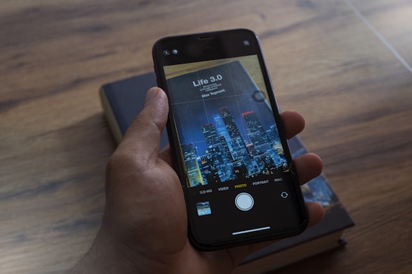 A hand is holding a smartphone, displaying a book cover image titled 'Life 3.0' by Max Tegmark, with a city skyline featured prominently. The background shows a wooden surface with a closed book.