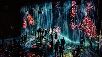 A dark room is filled with people viewing a digital art installation featuring cascading virtual waterfalls against an ethereal backdrop. The artwork is interspersed with vivid floral patterns, creating a vibrant and immersive environment.