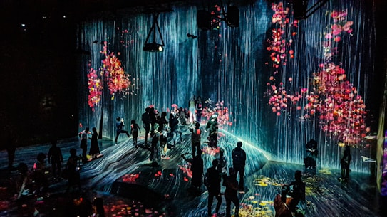 A dark room is filled with people viewing a digital art installation featuring cascading virtual waterfalls against an ethereal backdrop. The artwork is interspersed with vivid floral patterns, creating a vibrant and immersive environment.