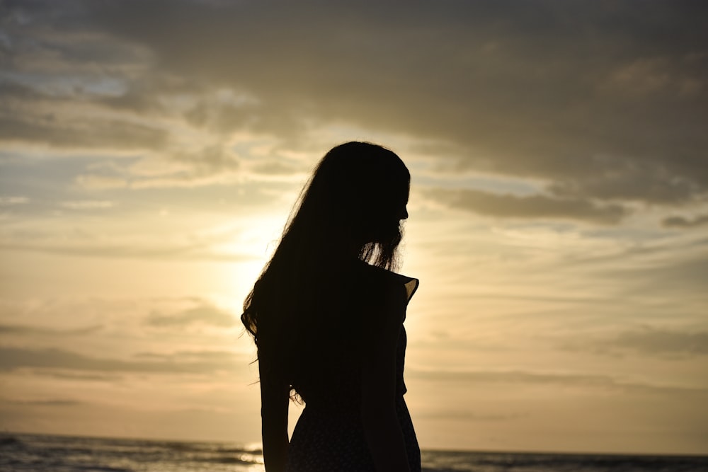 silhouette of woman standing near body of water during sunset