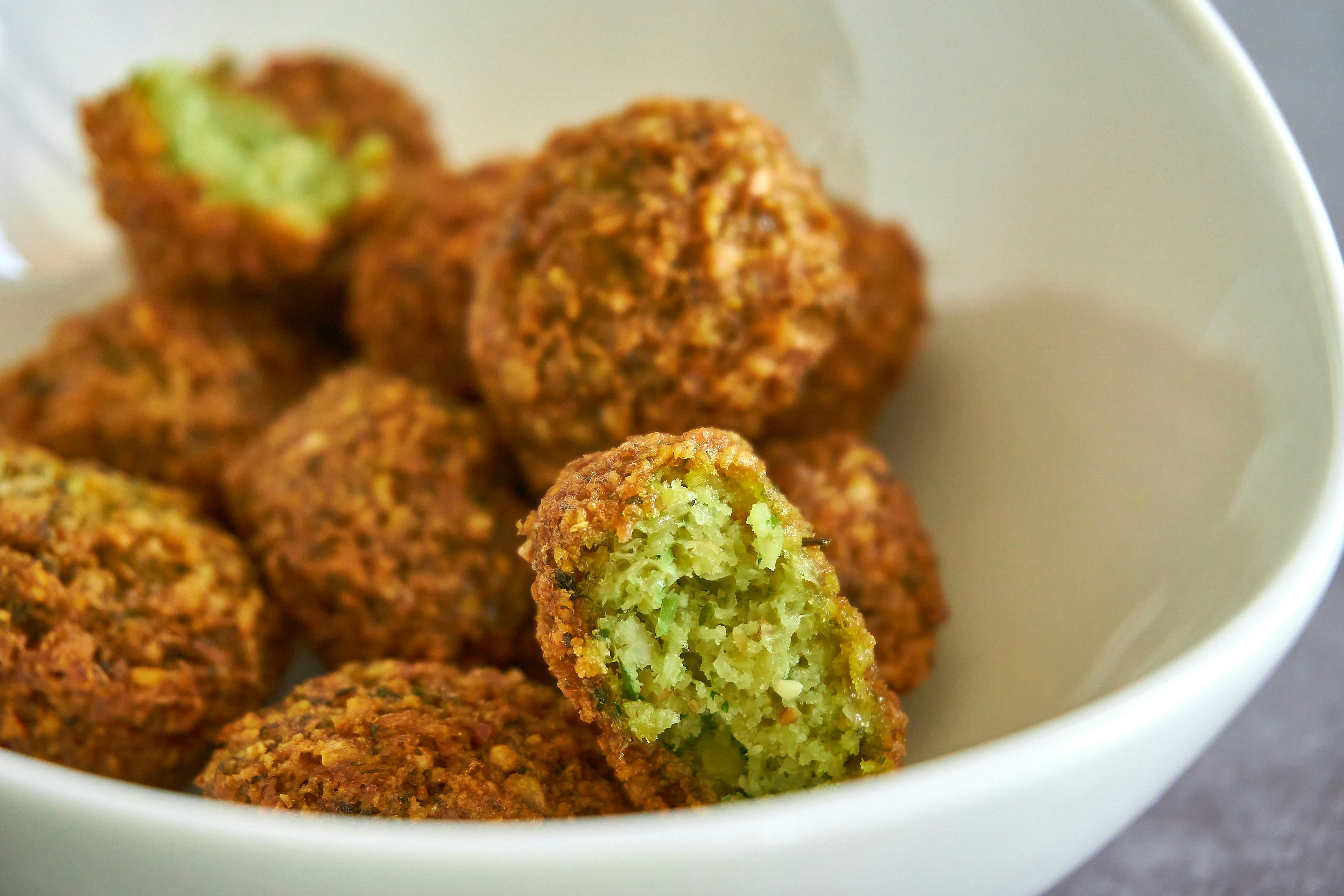 A bowl of falafels with one falafel revealing its green centre in a white bowl.