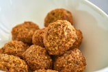A bowl filled with several pieces of golden-brown, crispy falafel. The surface texture of each piece appears crunchy and coarse, suggesting they have been deep-fried to achieve the crispy exterior. The bowl is white, providing a contrast to the brown color of the falafel.