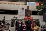 A person sits on top of a van holding a large protest sign that reads 'Truth is Not Justice, No Justice, No Peace'. Other individuals are visible nearby, one of whom is looking at a phone. T-shirts with protest slogans are displayed on the van. The setting appears to be urban with bicycles in the background and a partially visible building.