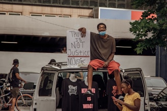 A person sits on top of a van holding a large protest sign that reads 'Truth is Not Justice, No Justice, No Peace'. Other individuals are visible nearby, one of whom is looking at a phone. T-shirts with protest slogans are displayed on the van. The setting appears to be urban with bicycles in the background and a partially visible building.