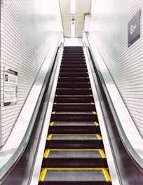 An escalator in a tiled interior space, with metal handrails and steps marked with yellow safety lines. The walls are covered with small white tiles and there are signs in Japanese, indicating a location in Japan.