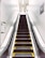 An escalator in a tiled interior space, with metal handrails and steps marked with yellow safety lines. The walls are covered with small white tiles and there are signs in Japanese, indicating a location in Japan.