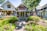 An inviting cottage-style house with intricate woodwork, surrounded by a lush garden filled with vibrant flowers and greenery. American flags are placed throughout the garden, enhancing the patriotic theme. Several other charming cottages are visible in the background, shaded by large, leafy trees.
