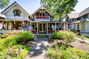 An inviting cottage-style house with intricate woodwork, surrounded by a lush garden filled with vibrant flowers and greenery. American flags are placed throughout the garden, enhancing the patriotic theme. Several other charming cottages are visible in the background, shaded by large, leafy trees.