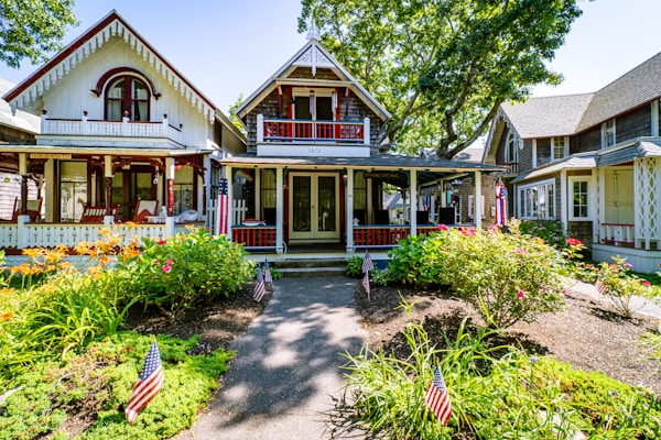 An inviting cottage-style house with intricate woodwork, surrounded by a lush garden filled with vibrant flowers and greenery. American flags are placed throughout the garden, enhancing the patriotic theme. Several other charming cottages are visible in the background, shaded by large, leafy trees.