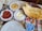 On a table, various dishes are arranged, including a bowl of thin flatbread with black seeds, a plate of creamy white dip drizzled with oil, a serving of red sauce or paste on a plate, and a bowl of hummus-like spread. There is a container of ayran, a traditional yogurt-based beverage, and a set of metal cutlery beside white plates.
