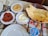 On a table, various dishes are arranged, including a bowl of thin flatbread with black seeds, a plate of creamy white dip drizzled with oil, a serving of red sauce or paste on a plate, and a bowl of hummus-like spread. There is a container of ayran, a traditional yogurt-based beverage, and a set of metal cutlery beside white plates.