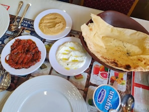 On a table, various dishes are arranged, including a bowl of thin flatbread with black seeds, a plate of creamy white dip drizzled with oil, a serving of red sauce or paste on a plate, and a bowl of hummus-like spread. There is a container of ayran, a traditional yogurt-based beverage, and a set of metal cutlery beside white plates.