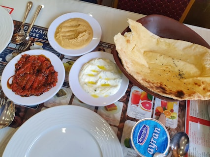 On a table, various dishes are arranged, including a bowl of thin flatbread with black seeds, a plate of creamy white dip drizzled with oil, a serving of red sauce or paste on a plate, and a bowl of hummus-like spread. There is a container of ayran, a traditional yogurt-based beverage, and a set of metal cutlery beside white plates.
