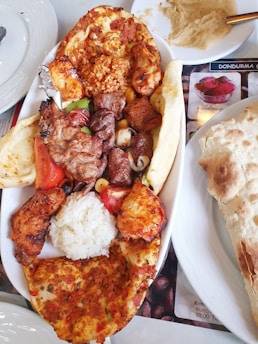 A platter filled with various types of grilled meats including chicken and lamb, accompanied by a scoop of rice, slices of flatbread, and vegetables. There is a separate plate with hummus and a portion of flatbread on the side.