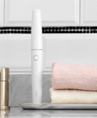 A sleek white electronic device with a circular button is placed horizontally near two folded towels, one pink and one cream. The device stands upright on a marble tray, with a metallic container nearby. The background features a tiled wall with white and black tiles.
