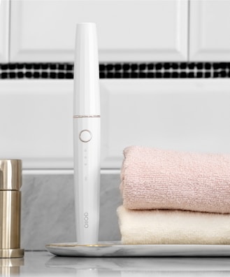 A sleek white electronic device with a circular button is placed horizontally near two folded towels, one pink and one cream. The device stands upright on a marble tray, with a metallic container nearby. The background features a tiled wall with white and black tiles.