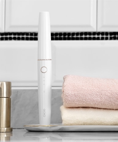 A sleek white electronic device with a circular button is placed horizontally near two folded towels, one pink and one cream. The device stands upright on a marble tray, with a metallic container nearby. The background features a tiled wall with white and black tiles.