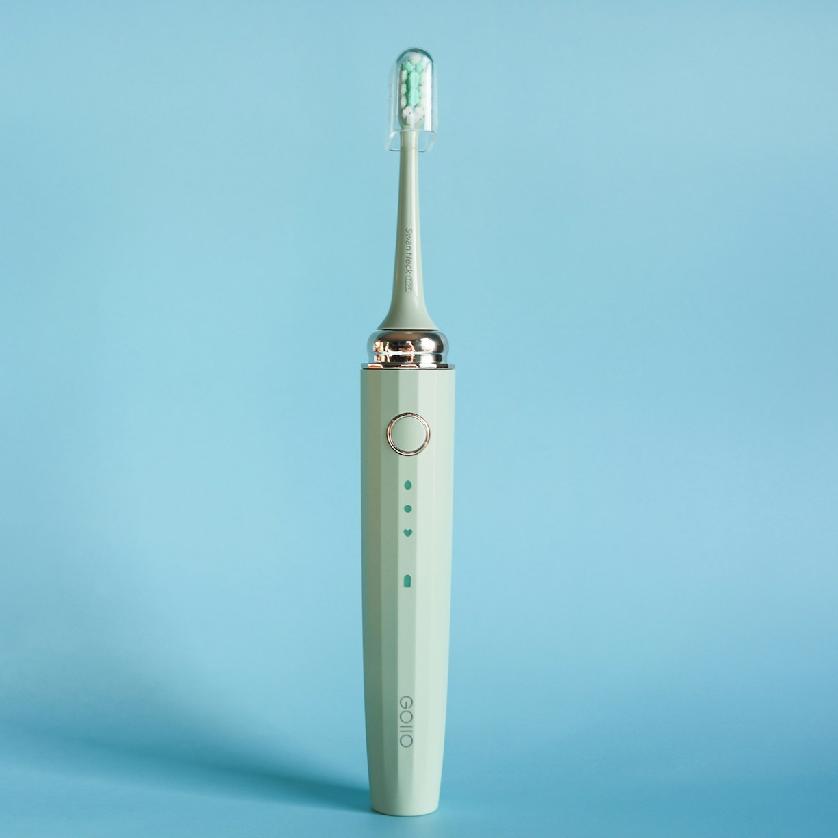 An electric toothbrush is standing upright against a solid light blue background. The toothbrush is a muted green color with a sleek design, featuring a silver ring near the top and an angled brush head with white and green bristles enclosed in a transparent cover.