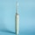 An electric toothbrush is standing upright against a solid light blue background. The toothbrush is a muted green color with a sleek design, featuring a silver ring near the top and an angled brush head with white and green bristles enclosed in a transparent cover.