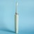 An electric toothbrush is standing upright against a solid light blue background. The toothbrush is a muted green color with a sleek design, featuring a silver ring near the top and an angled brush head with white and green bristles enclosed in a transparent cover.