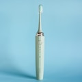 An electric toothbrush is standing upright against a solid light blue background. The toothbrush is a muted green color with a sleek design, featuring a silver ring near the top and an angled brush head with white and green bristles enclosed in a transparent cover.