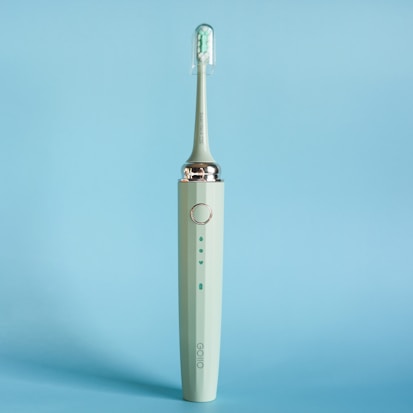 An electric toothbrush is standing upright against a solid light blue background. The toothbrush is a muted green color with a sleek design, featuring a silver ring near the top and an angled brush head with white and green bristles enclosed in a transparent cover.