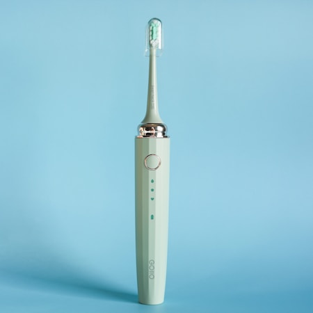 An electric toothbrush is standing upright against a solid light blue background. The toothbrush is a muted green color with a sleek design, featuring a silver ring near the top and an angled brush head with white and green bristles enclosed in a transparent cover.