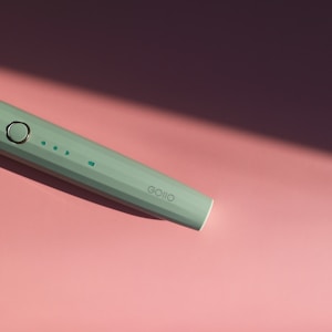 A mint green electronic device with minimalist design lays diagonally on a pink surface. The device features a round button and small indicator lights, suggesting functionality. The lighting creates a soft shadow, adding depth to the composition.
