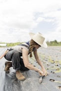 A person wearing a wide-brimmed hat and sunglasses is kneeling on a farm field, planting seedlings. They are dressed in work attire, including boots and overalls. The field has rows of small plants with holes prepared for planting, and the sky is partly cloudy.